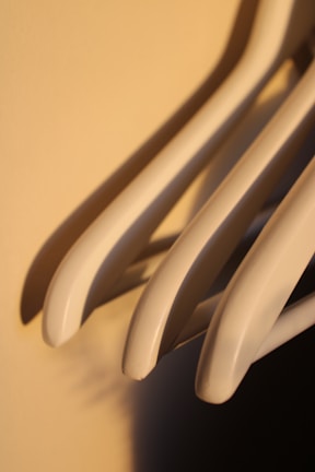 A close-up image of several light-colored plastic clothes hangers casting shadows on a warm-colored wall. The hangers are positioned in a slightly diagonal orientation, with soft lighting highlighting their curves.