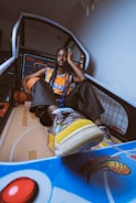 A person with long braided hair sits casually inside a basketball arcade game. They are wearing a sports jersey, black pants, and colorful sneakers. There is a basketball next to them, and the arcade's vibrant buttons and scoreboard are visible.