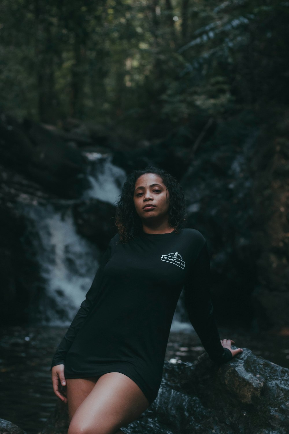 a woman in a black dress sitting on a rock in front of a waterfall