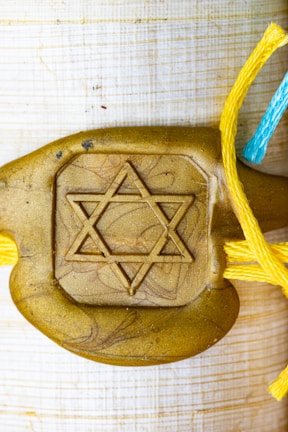 A gold-colored decorative piece features a prominent Star of David symbol in the center. It is attached to strings of different colors: yellow and blue, suggesting a handmade or artistic element.
