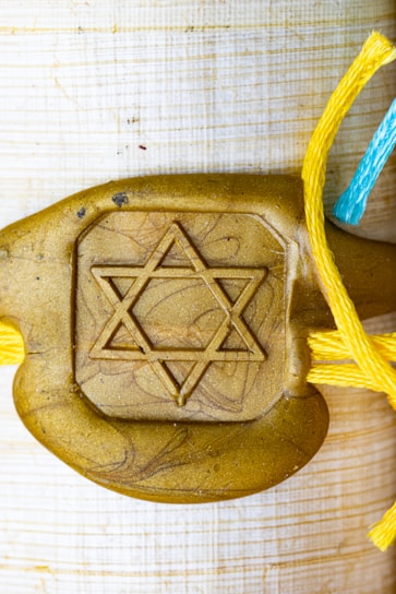 A gold-colored decorative piece features a prominent Star of David symbol in the center. It is attached to strings of different colors: yellow and blue, suggesting a handmade or artistic element.