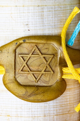 A gold-colored decorative piece features a prominent Star of David symbol in the center. It is attached to strings of different colors: yellow and blue, suggesting a handmade or artistic element.