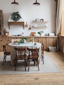 A cozy kitchen with wooden cabinetry and an organized wooden table surrounded by four chairs. The wall features open shelves with plates, cups, and various kitchen items. Potted plants on the table and counter add a touch of greenery. The room is brightly lit by natural light streaming in through a large window, with a soft beige rug under the table.