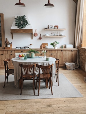 A cozy kitchen with wooden cabinetry and an organized wooden table surrounded by four chairs. The wall features open shelves with plates, cups, and various kitchen items. Potted plants on the table and counter add a touch of greenery. The room is brightly lit by natural light streaming in through a large window, with a soft beige rug under the table.