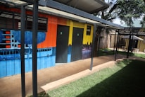 A colorful wall mural depicting urban cityscape imagery, including silhouettes of buildings and trees. The mural is painted on a metal structure that appears to be a restroom facility, complete with doors marked for use. The setting includes a green lawn and a shaded seating area with tables and chairs under an umbrella.