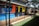 A colorful wall mural depicting urban cityscape imagery, including silhouettes of buildings and trees. The mural is painted on a metal structure that appears to be a restroom facility, complete with doors marked for use. The setting includes a green lawn and a shaded seating area with tables and chairs under an umbrella.