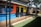 A colorful wall mural depicting urban cityscape imagery, including silhouettes of buildings and trees. The mural is painted on a metal structure that appears to be a restroom facility, complete with doors marked for use. The setting includes a green lawn and a shaded seating area with tables and chairs under an umbrella.