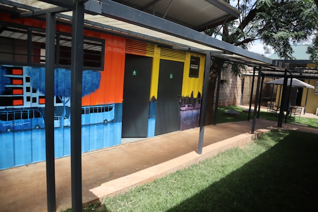 A colorful wall mural depicting urban cityscape imagery, including silhouettes of buildings and trees. The mural is painted on a metal structure that appears to be a restroom facility, complete with doors marked for use. The setting includes a green lawn and a shaded seating area with tables and chairs under an umbrella.