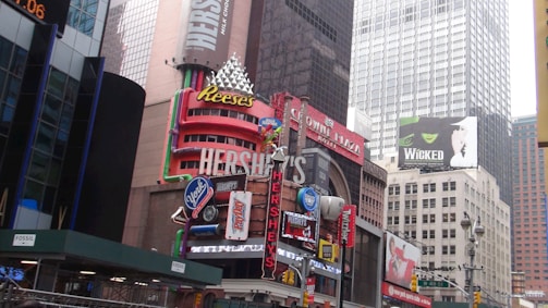 A busy urban scene featuring numerous colorful and prominent advertisements for products like Reese's and Hershey's. Tall buildings with large billboards, including one for the musical 'Wicked', dominate the background. The scene includes other brand signs and logos, all contributing to a lively city atmosphere.