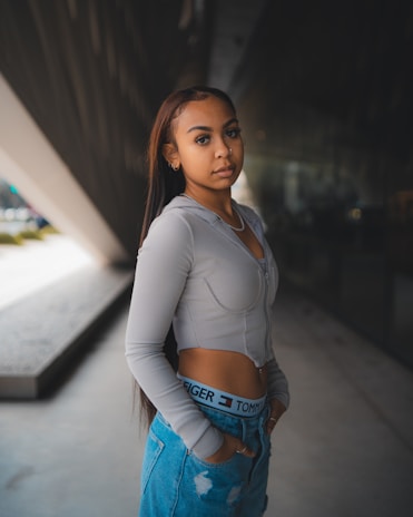 A young woman stands confidently in an urban setting. She is wearing a long-sleeve gray top and casual blue jeans with designer branding visible on the waistband. Her long hair flows down her shoulders and she gazes directly at the camera with a calm expression. The background features modern architecture with a large triangular structure and reflective glass.