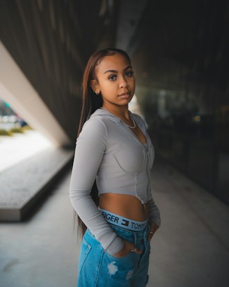 A young woman stands confidently in an urban setting. She is wearing a long-sleeve gray top and casual blue jeans with designer branding visible on the waistband. Her long hair flows down her shoulders and she gazes directly at the camera with a calm expression. The background features modern architecture with a large triangular structure and reflective glass.