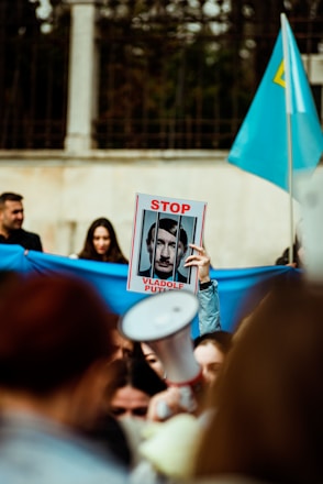A group of people appears to be at a protest or demonstration. One person is holding a placard with the words 'Stop Vladolf Putin' and an image of a man behind bars. There is a blue flag waving in the background, and a megaphone is visible in the crowd.