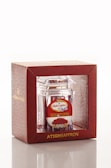 A product packaging featuring a clear container with saffron inside, encased in a decorative red box. The box has gold lettering and a patterned design, with branding and additional text in another language on a label.
