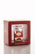 A product packaging featuring a clear container with saffron inside, encased in a decorative red box. The box has gold lettering and a patterned design, with branding and additional text in another language on a label.