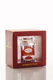 A product packaging featuring a clear container with saffron inside, encased in a decorative red box. The box has gold lettering and a patterned design, with branding and additional text in another language on a label.