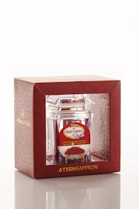A product packaging featuring a clear container with saffron inside, encased in a decorative red box. The box has gold lettering and a patterned design, with branding and additional text in another language on a label.