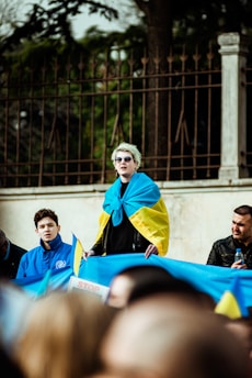 A group of people are gathered outdoors, participating in a protest or demonstration. One individual stands prominently, wrapped in a flag with blue and yellow colors, likely representing the Ukrainian flag. The person is wearing sunglasses and addressing the crowd. Others hold signs and flags with similar colors. In the background, there is a fence and trees, indicating an open area or park setting.