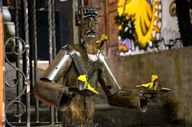 A metal sculpture resembling a humanoid figure is situated outdoors. It appears to be made from pipes and scrap metal, with a slightly abstract and rustic design. The sculpture is holding small yellow birds made from the same metal material. In the background, there is a colorful mural partially visible, enhancing the artistic setting.