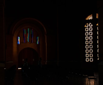 A dimly lit interior space showcases colorful stained glass windows with abstract designs. The walls are adorned with intricate patterns allowing soft light to filter through, creating a serene and contemplative atmosphere.
