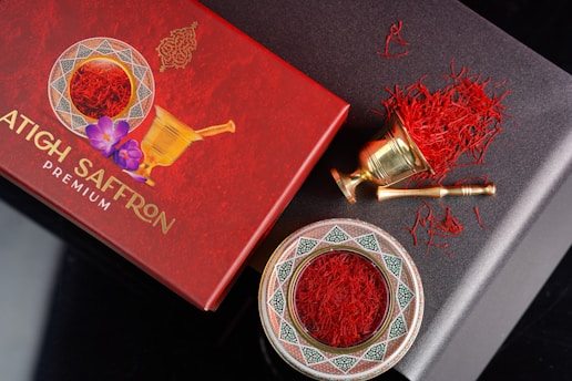 An elegant arrangement of saffron threads is displayed with a decorative red box and a traditional brass mortar and pestle. The box, labeled 'Premium Saffron,' features intricate designs and a floral element. The saffron is also presented in an ornate dish with geometric patterns.