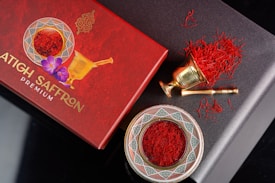 An elegant arrangement of saffron threads is displayed with a decorative red box and a traditional brass mortar and pestle. The box, labeled 'Premium Saffron,' features intricate designs and a floral element. The saffron is also presented in an ornate dish with geometric patterns.