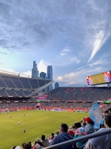 A sports stadium filled with spectators watching a soccer match. Players are on the field in action, and a large scoreboard displays information about the game. The stadium has blue seating and is surrounded by tall city buildings visible in the background against a partly cloudy sky. Fans are dressed casually, some waving flags and wearing team colors.