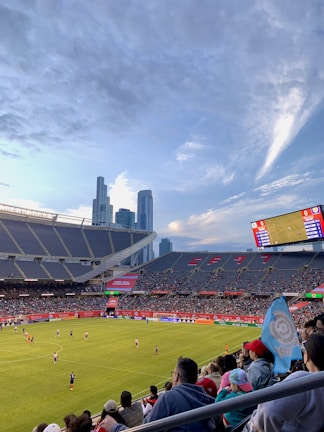 A sports stadium filled with spectators watching a soccer match. Players are on the field in action, and a large scoreboard displays information about the game. The stadium has blue seating and is surrounded by tall city buildings visible in the background against a partly cloudy sky. Fans are dressed casually, some waving flags and wearing team colors.