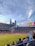 A sports stadium filled with spectators watching a soccer match. Players are on the field in action, and a large scoreboard displays information about the game. The stadium has blue seating and is surrounded by tall city buildings visible in the background against a partly cloudy sky. Fans are dressed casually, some waving flags and wearing team colors.