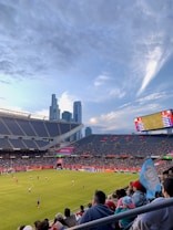 A sports stadium filled with spectators watching a soccer match. Players are on the field in action, and a large scoreboard displays information about the game. The stadium has blue seating and is surrounded by tall city buildings visible in the background against a partly cloudy sky. Fans are dressed casually, some waving flags and wearing team colors.