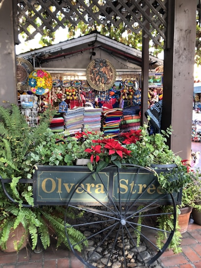 A vibrant market stall with colorful woven textiles and decorative plates on display. The stall is surrounded by lush green plants and bright red flowers. A wooden cart with 'Olvera Street' painted on the side is prominently featured in front of the stall, adding a rustic charm to the scene. Hanging decorations, including intricate and colorful circular designs, enhance the market atmosphere.