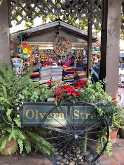 A vibrant market stall with colorful woven textiles and decorative plates on display. The stall is surrounded by lush green plants and bright red flowers. A wooden cart with 'Olvera Street' painted on the side is prominently featured in front of the stall, adding a rustic charm to the scene. Hanging decorations, including intricate and colorful circular designs, enhance the market atmosphere.