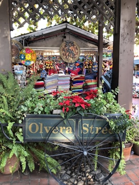 A vibrant market stall with colorful woven textiles and decorative plates on display. The stall is surrounded by lush green plants and bright red flowers. A wooden cart with 'Olvera Street' painted on the side is prominently featured in front of the stall, adding a rustic charm to the scene. Hanging decorations, including intricate and colorful circular designs, enhance the market atmosphere.
