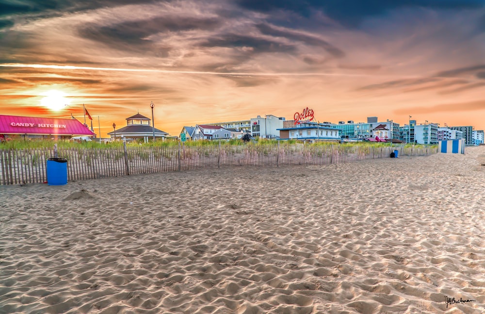 a sandy beach with a fence and buildings in the background