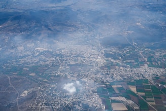 An aerial view of a sprawling cityscape interspersed with patches of agricultural fields. The landscape is surrounded by a range of mountains, partially obscured by haze. Various geometric patterns are formed by the fields, and a cloud is visible floating over the city area.