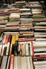 A large collection of assorted books is neatly arranged, mostly spine-up and lying on their sides. The books vary in color, size, and thickness, suggesting a diverse selection of genres and topics.