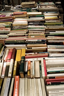 A large collection of assorted books is neatly arranged, mostly spine-up and lying on their sides. The books vary in color, size, and thickness, suggesting a diverse selection of genres and topics.