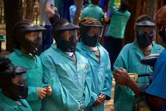 A group of individuals wearing teal overalls and protective masks stand together. They appear to be preparing for a paintball game, as suggested by the masks and a paintball gun in view. The setting is outdoors with trees and other people in the background, some dressed similarly.