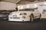 A glossy white sports car is on display at a car show, surrounded by other modified vehicles. The car features sleek design elements like aerodynamic bodywork and stylish rims, showcased under a tent with bright overhead lighting. The setting suggests a professional automotive exhibition.