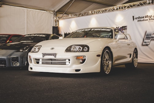 A glossy white sports car is on display at a car show, surrounded by other modified vehicles. The car features sleek design elements like aerodynamic bodywork and stylish rims, showcased under a tent with bright overhead lighting. The setting suggests a professional automotive exhibition.