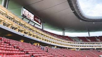 A large, modern stadium with red seats and a sleek, expansive roof structure. A few people are scattered among the seats, and there are large digital screens displaying advertisements. The setting is well-lit with multiple levels visible, suggesting a multi-tiered structure.