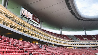 A large, modern stadium with red seats and a sleek, expansive roof structure. A few people are scattered among the seats, and there are large digital screens displaying advertisements. The setting is well-lit with multiple levels visible, suggesting a multi-tiered structure.