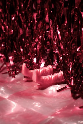 Pink metallic streamers hang down, partially covering a pair of pink high-heeled boots. The scene is set against a shiny, reflective surface that enhances the pink hues throughout the image.