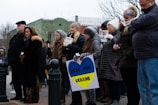 A group of people stand together outdoors, holding signs with messages such as 'Pray for Ukraine' and 'Stop War'. They are dressed in warm clothing, suggesting cold weather. The expressions on their faces seem solemn and serious.
