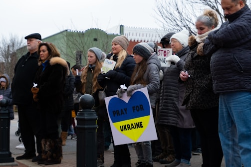 A group of people stand together outdoors, holding signs with messages such as 'Pray for Ukraine' and 'Stop War'. They are dressed in warm clothing, suggesting cold weather. The expressions on their faces seem solemn and serious.