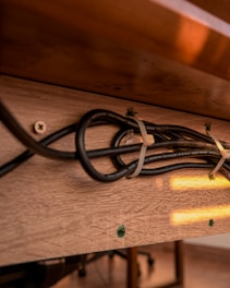A wooden surface with a cluster of black cables fastened by cable ties. The cables are secured against the brown wooden texture with screws partially visible. Warm light reflects off the surface, casting a soft glow. The background shows part of a floor and furniture legs.