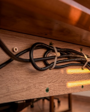 A wooden surface with a cluster of black cables fastened by cable ties. The cables are secured against the brown wooden texture with screws partially visible. Warm light reflects off the surface, casting a soft glow. The background shows part of a floor and furniture legs.
