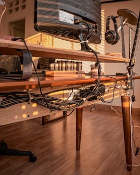 A home office setup features a computer monitor on a wooden desk. Underneath the desk, numerous cables are neatly organized and bundled together. On the desk are various office items including a set of notebooks, a desk lamp, and headphones hung on a stand. The room is warmly lit with natural sunlight streaming through window blinds, creating a pattern of light and shadow on the desk and floor.