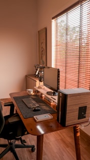 A cozy and minimalist home office setup featuring a wooden desk with a computer monitor, keyboard, and mouse. A desktop tower is positioned beside the monitor. A smartphone is placed on the desk, alongside a white mug. Light filters through wooden blinds, casting warm patterns on the interior. The overall ambiance is clean and organized, with a soft glow of natural light.