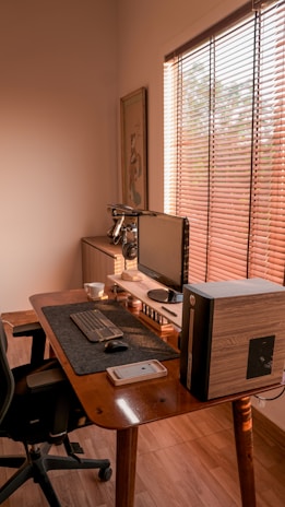 A cozy and minimalist home office setup featuring a wooden desk with a computer monitor, keyboard, and mouse. A desktop tower is positioned beside the monitor. A smartphone is placed on the desk, alongside a white mug. Light filters through wooden blinds, casting warm patterns on the interior. The overall ambiance is clean and organized, with a soft glow of natural light.