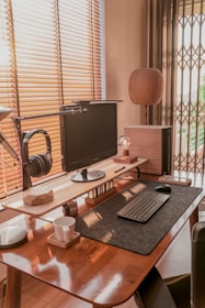 A neatly organized home office setup with a modern aesthetic. The desk features a computer monitor mounted on a raised stand, a keyboard and mouse placed on a felt mat, and a pair of black headphones hanging from a side mount. There is a cup on a coaster, a small lamp, and a decorative wooden object on the desk. Natural light filters through the wooden blinds, illuminating the space with a warm glow.